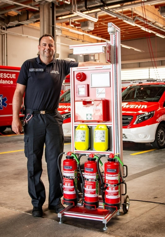 Ein Feuerwehrmann steht neben einem Feuerwehr Rollcontainer für Erste Hilfe vor einem Löschfahrzeug auf dem Gelände der Feuerwehrwache.